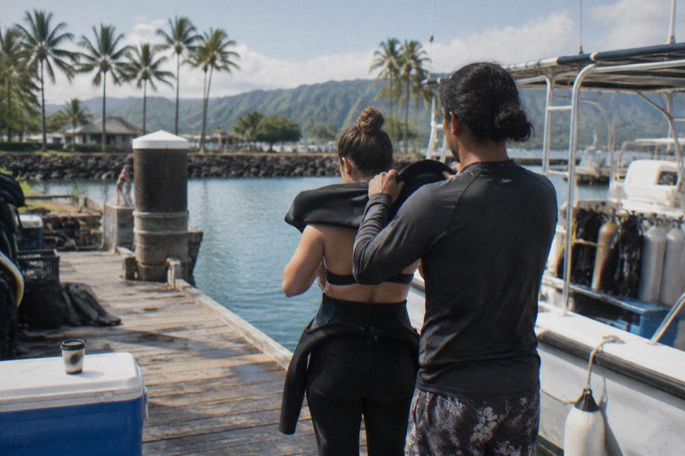 Crew member handing equipment to guests beside a boat at a wooden dock